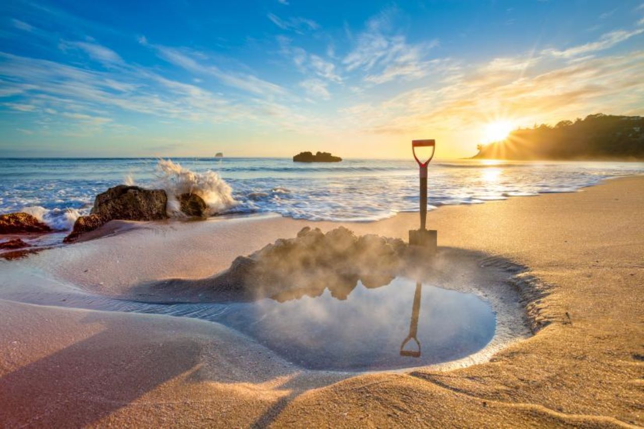 A small pool with steaming water on a beach with the sea and clear sky in the background